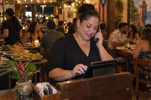 La anfitriona de un restaurante hablando por teléfono en la entrada, con el comedor lleno detrás.
